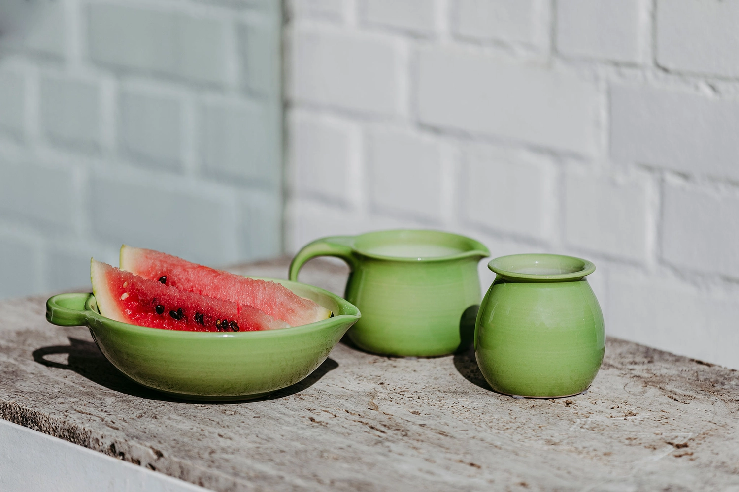 Keramik Atelier Melis - Töpferkurse - Auf einem rustikalen Holztisch steht neben einem kleinen grünen Krug und einem grünen Topf eine hellgrüne Schüssel mit Wassermelonenscheiben; im Hintergrund ist eine weiße Backsteinmauer zu sehen.