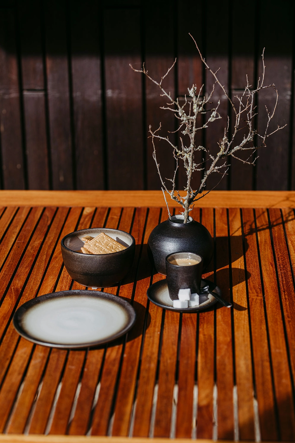 Keramik Atelier Melis - Töpferkurse - Nahaufnahme eines mit dunklen Keramiktellern, einer Tasse Kaffee mit Zuckerwürfeln und einer Vase mit trockenen Zweigen gedeckten Holzlattentisches.