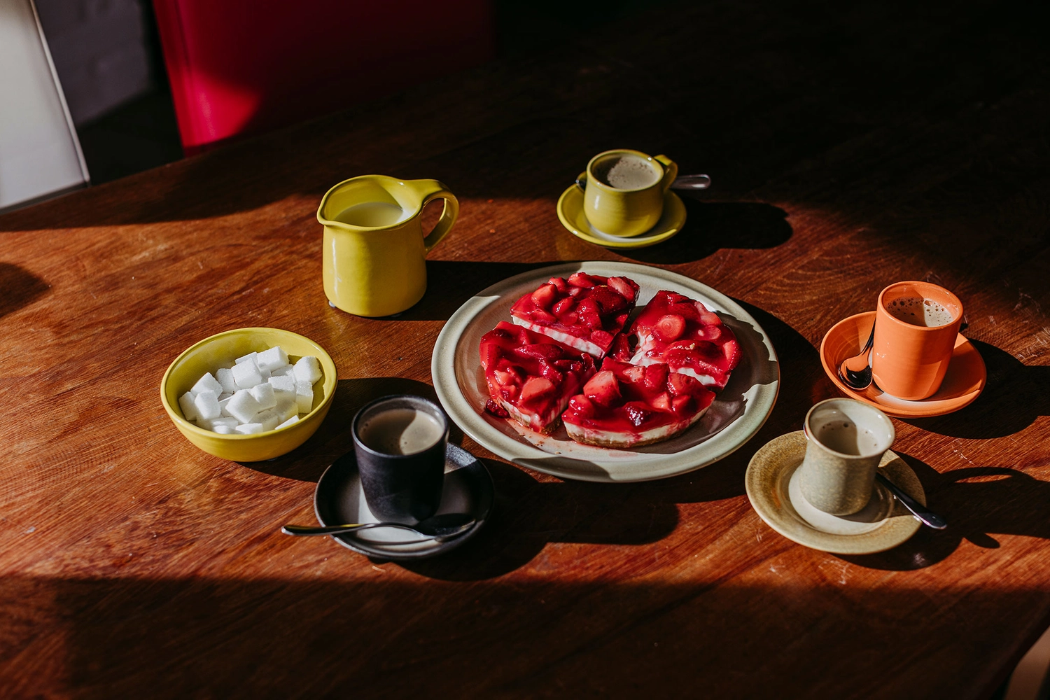 Keramik Atelier Melis - Töpferkurse - Ein Holztisch, gedeckt mit gelben und orangefarbenen Keramiktassen, einem Sahnekännchen, einer Schale mit Zuckerwürfeln und einem Teller mit Erdbeertörtchenstücken.