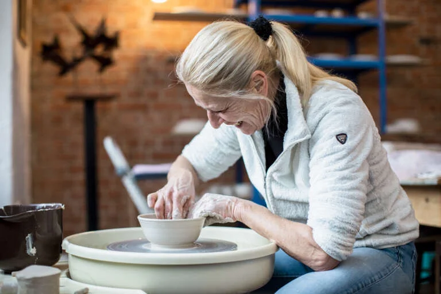 Keramik Atelier Melis - Töpferkurse - Eine Frau mit blonden Haaren in einer weißen Fleecejacke arbeitet an einer Töpferscheibe mit Ton.