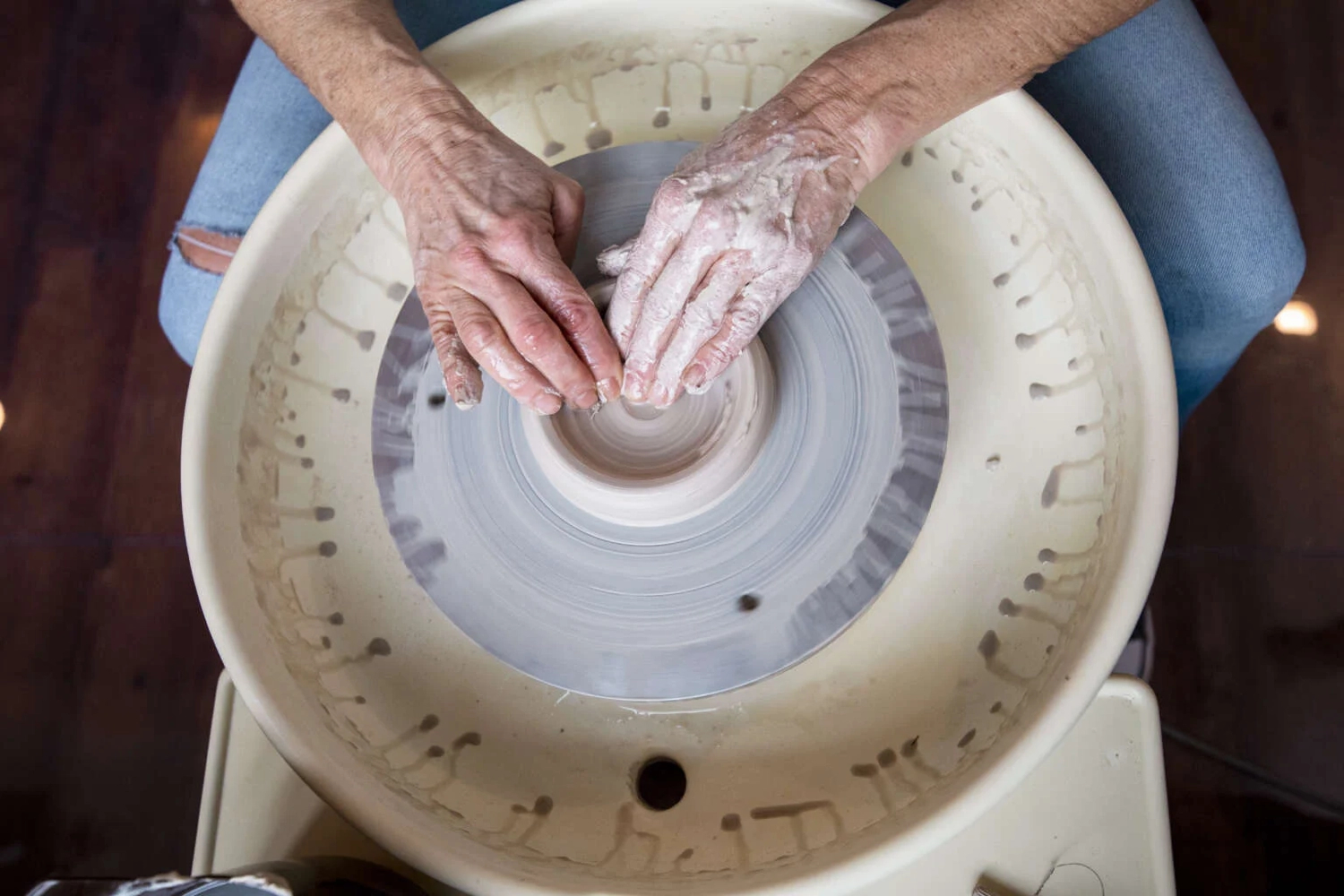 Keramik Atelier Melis - Töpferkurse - Nahaufnahme aus der Vogelperspektive: Eine Person formt mit nassen Händen einen Tonhaufen auf einer sich drehenden Töpferscheibe.