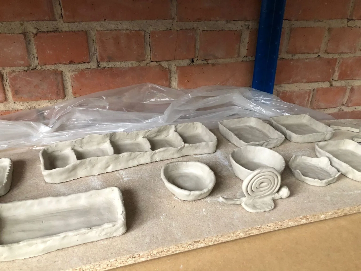 Keramik Atelier Melis - Töpferkurse - Eine Sammlung unfertiger, grauer Tonkeramikstücke, darunter kleine Schalen, rechteckige Tabletts und eine gewickelte Tonrose, die auf einer Holzfläche vor einer Backsteinmauer stehen.