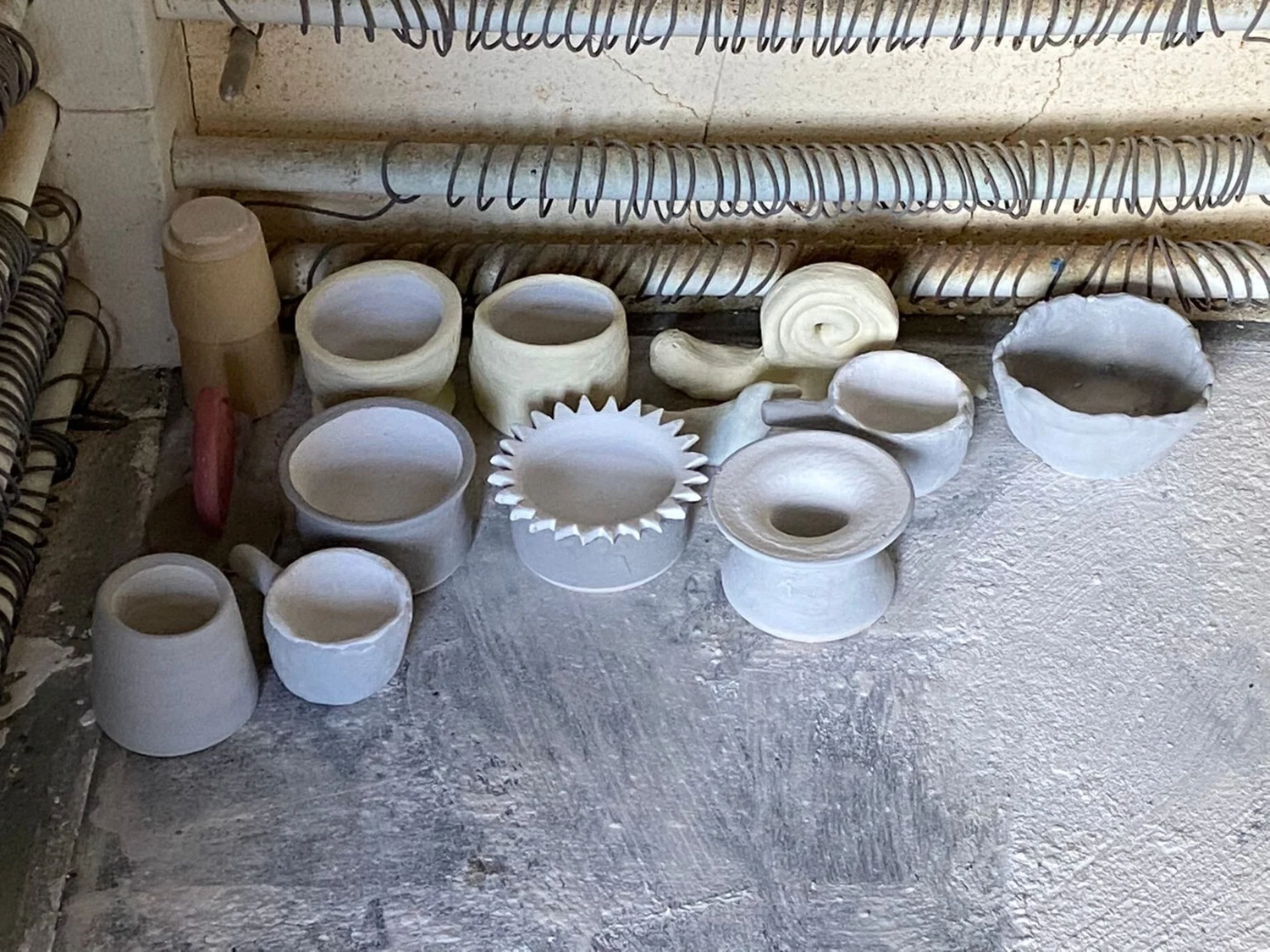 Keramik Atelier Melis - Töpferkurse - Eine Ansammlung unfertiger, weißer Keramikschalen, -tassen und -töpfe in verschiedenen Formen und Größen steht auf einer grauen, strukturierten Oberfläche, vermutlich in einer Töpferei oder in der Nähe eines Brennofens. Im Hintergrund sind Heizspiralen zu erkennen.