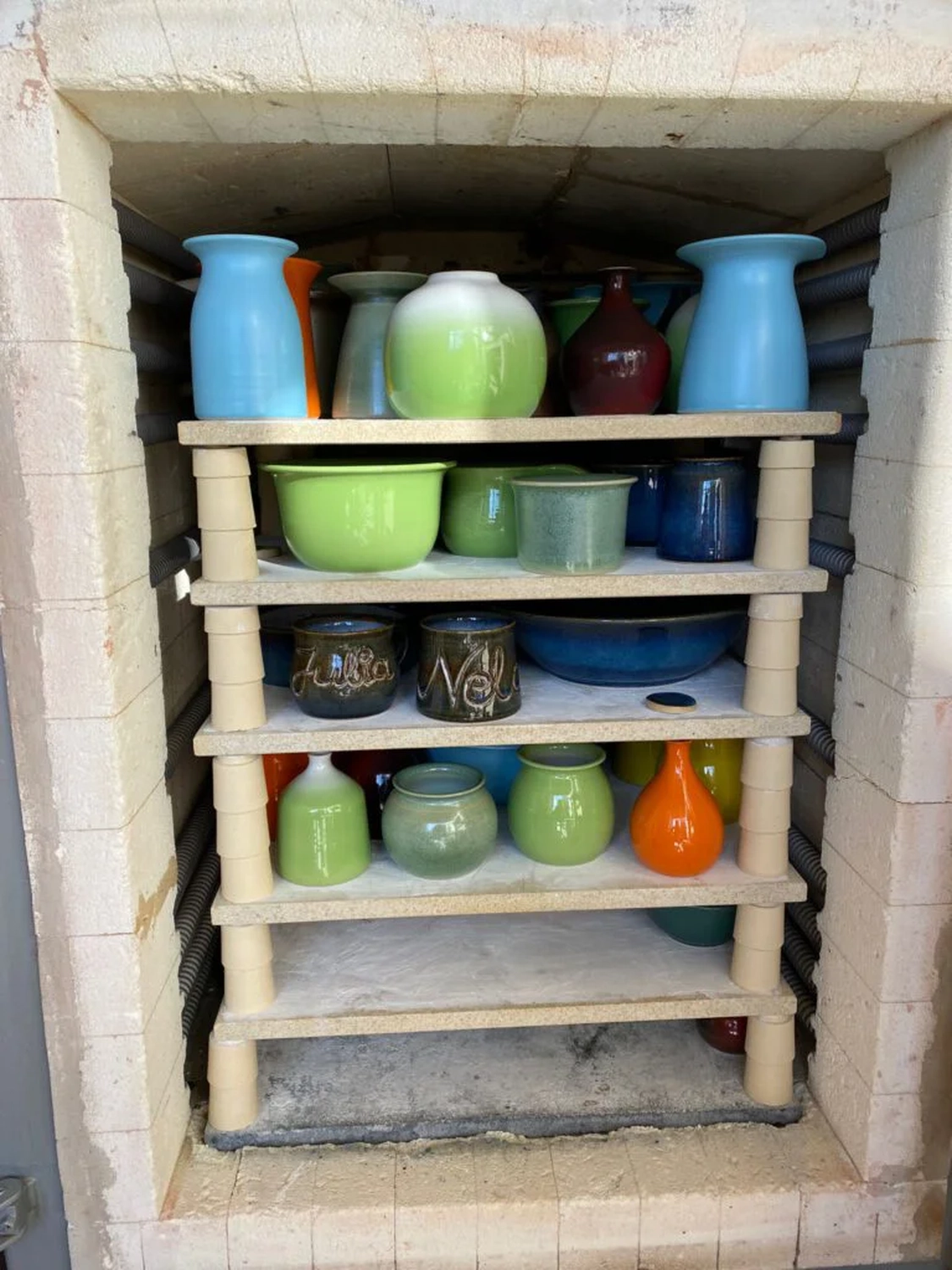 Keramik Atelier Melis - Töpferkurse - Ein mehrstöckiges Holzregal, gefüllt mit verschiedenen glasierten Keramiktöpfen, Vasen und Schalen, befindet sich in einem Brennofen.