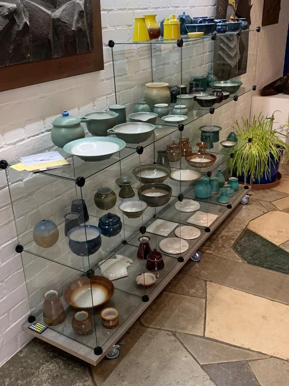 Keramik Atelier Melis - Töpferkurse - Eine Glasvitrine ist gefüllt mit verschiedenen Keramikwaren in gedeckten Grün-, Blau- und Brauntönen. Rechts daneben steht eine große grüne Zimmerpflanze in einem blauen Topf.