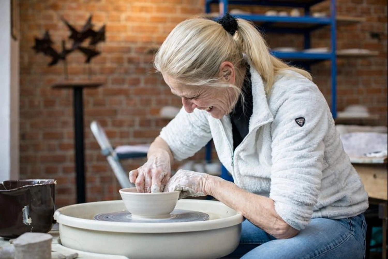 Keramik Atelier Melis - Töpferkurse - Eine Frau mit blonden Haaren in einer weißen Fleecejacke arbeitet an einer Töpferscheibe mit Ton.