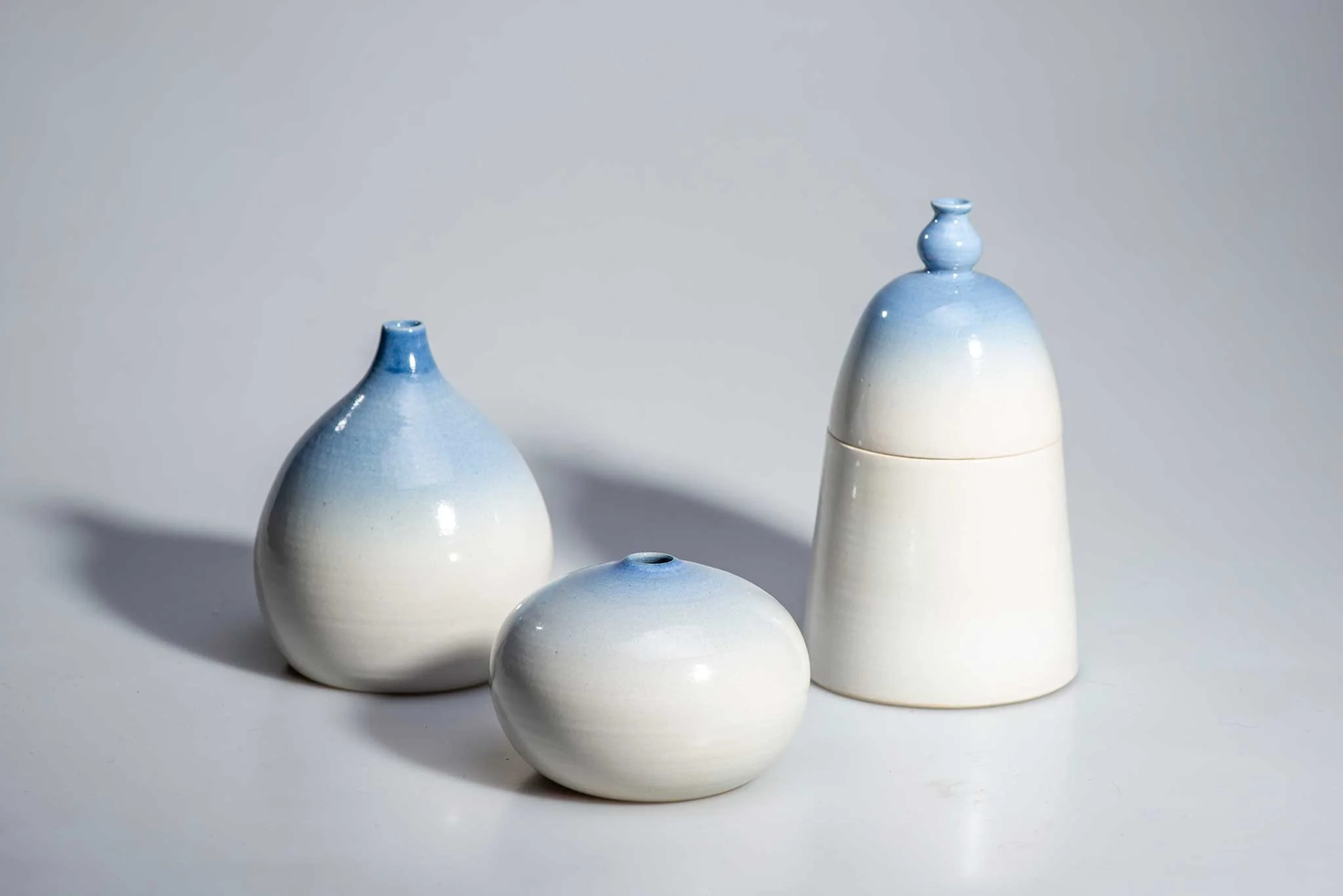 Keramik Atelier Melis - Töpferkurse - Drei Keramikobjekte mit einer von Weiß nach Blau verlaufenden Glasur, darunter eine tropfenförmige Vase, eine kleine runde Vase und ein Behälter mit Deckel.