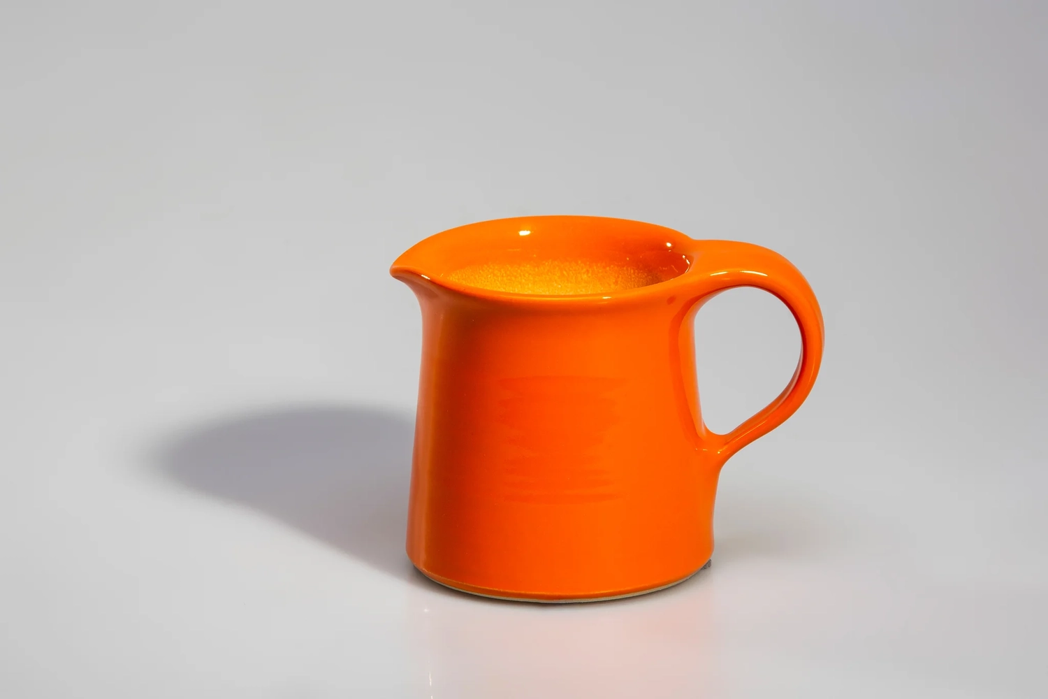 Keramik Atelier Melis - Töpferkurse - Ein kleiner, glänzender orangefarbener Keramikkrug, identifiziert als Bauer Vintage Plain Ware Pitcher, gefüllt mit einer gelblichen Flüssigkeit.