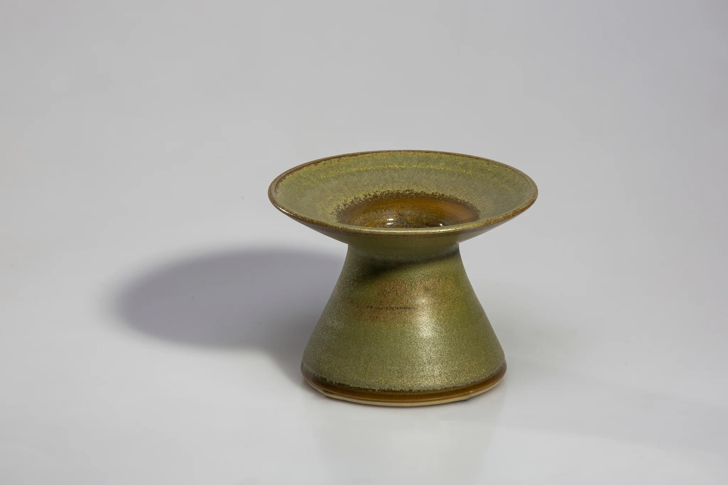 Keramik Atelier Melis - Töpferkurse - Ein antiker Kerzenhalter aus Keramik mit breitem, ausgestelltem Rand und Sockel, der mit einer gesprenkelten grünen und braunen Glasur versehen ist.