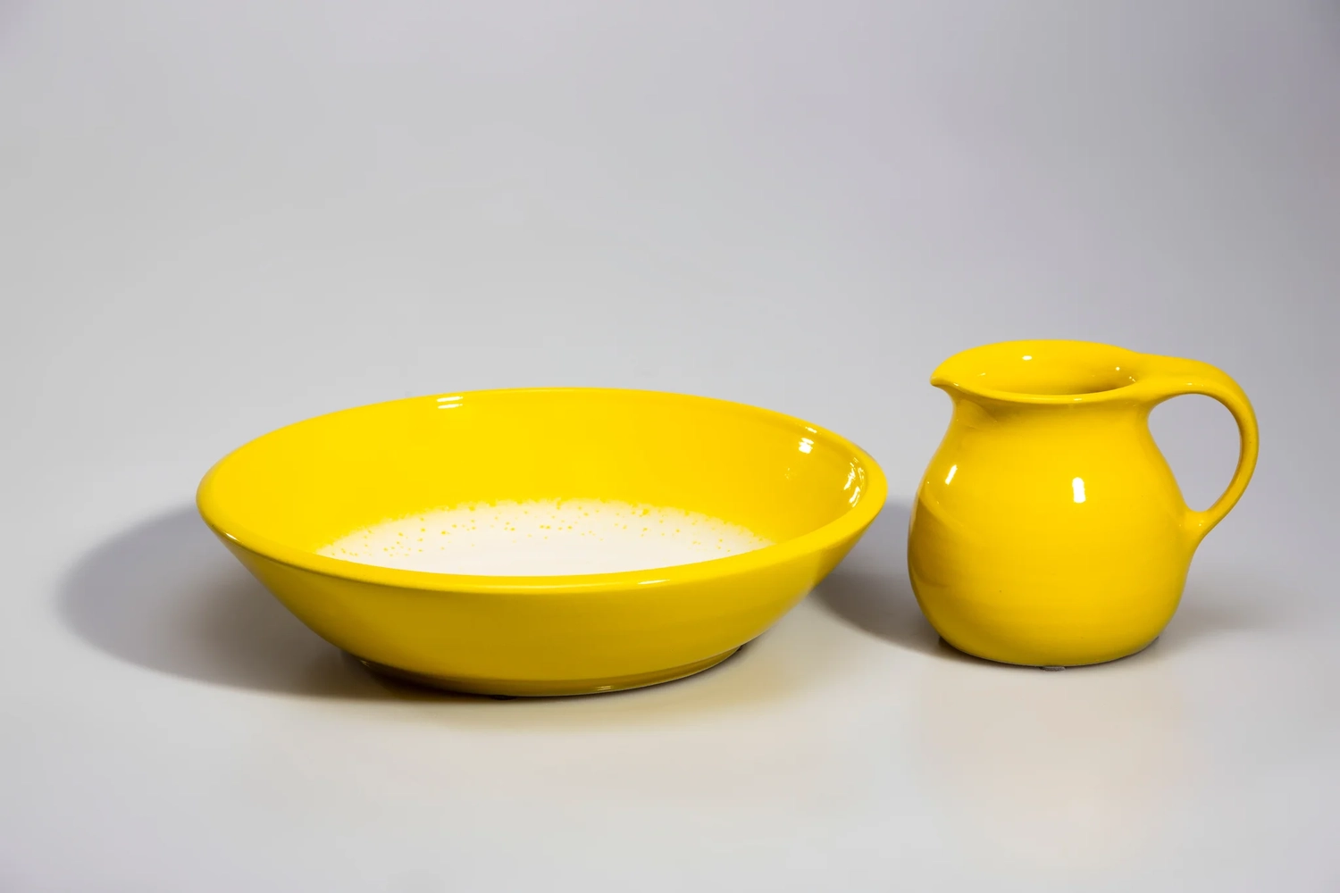 Keramik Atelier Melis - Töpferkurse - Eine leuchtend gelbe Keramikschale, gefüllt mit einer weißen Flüssigkeit, und ein kleiner, dazu passender gelber Krug auf grauem Untergrund.