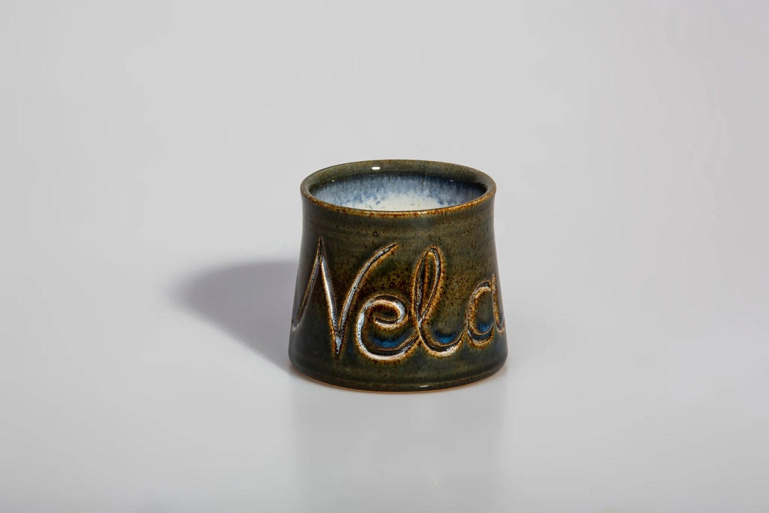 Keramik Atelier Melis - Töpferkurse - Ein kleiner, runder Keramiktopf mit dunkelgrüner und brauner Glasur. Das Wort „Vela“ ist in Schreibschrift in die Seite eingraviert und gibt den Blick auf eine hellere Farbe darunter frei. Der Rand ist blau-weiß glasiert.