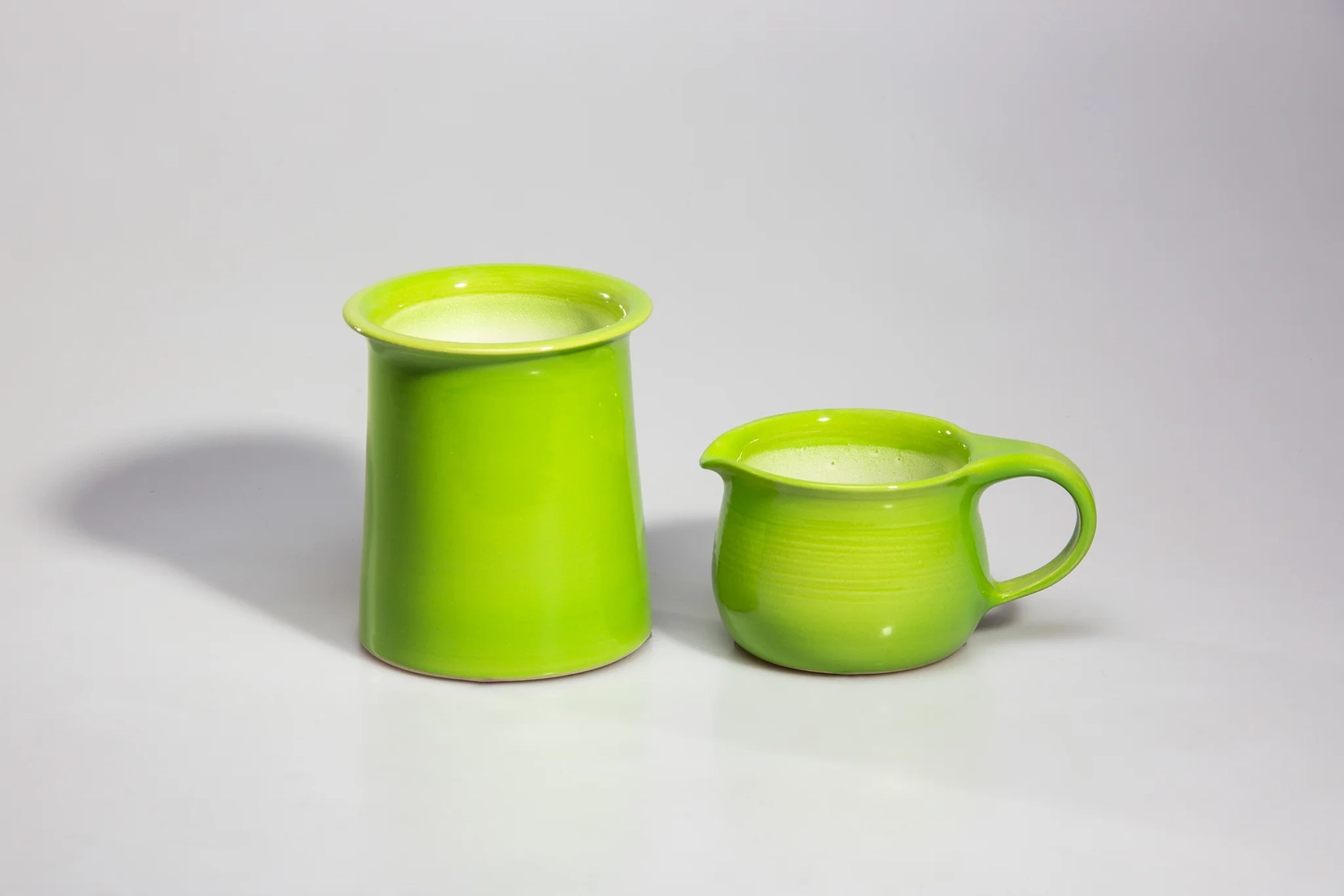 Keramik Atelier Melis - Töpferkurse - Zwei avocadogrüne Keramikgefäße, eines hoch und zylindrisch, das andere ein kleiner Krug, gefüllt mit einer weißen Flüssigkeit.