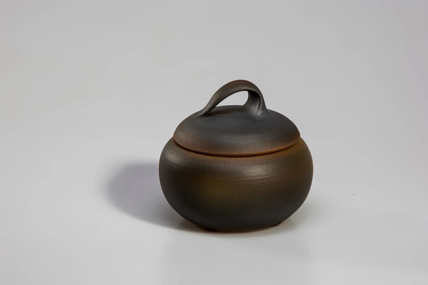 Keramik Atelier Melis - Töpferkurse - Ein kleiner, runder, dunkelbraun-schwarzer traditioneller japanischer Reistopf mit Deckel und Schlaufengriff.