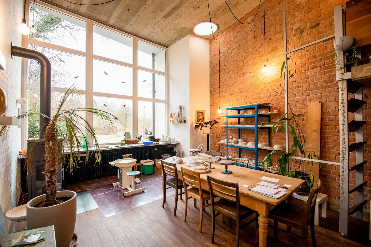 Keramik Atelier Melis - Töpferkurse - Ein helles, geräumiges Töpferatelier mit einem langen Holztisch, der mit Platzsets und Werkzeugen gedeckt ist, einer großen Fensterfront mit Blick ins Freie, freiliegenden Ziegelwänden, Holzboden, einer großen Topfpalme, einem blauen Metallregal mit Töpferwaren und einer Töpferscheibe in der Nähe des Fensters.