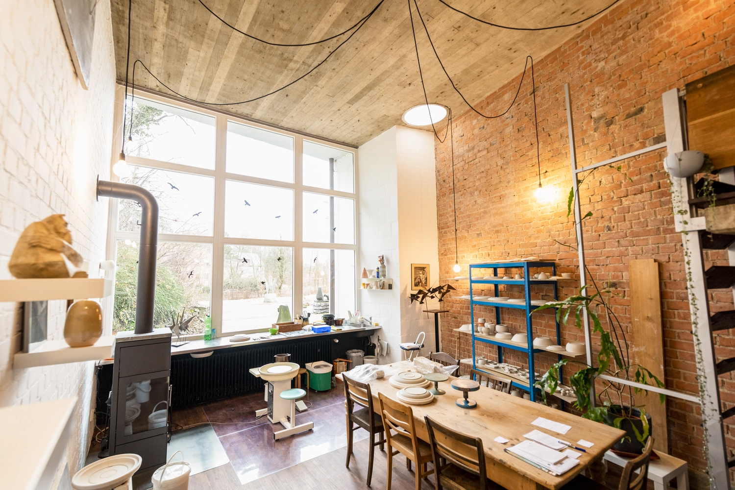 Keramik Atelier Melis - Töpferkurse - Ein helles, geräumiges Töpferatelier mit einem langen Holztisch, der mit Platzsets und Werkzeugen gedeckt ist, einer großen Fensterfront mit Blick ins Freie, freiliegenden Ziegelwänden, Holzboden, einer großen Topfpalme, einem blauen Metallregal mit Töpferwaren und einer Töpferscheibe in der Nähe des Fensters.