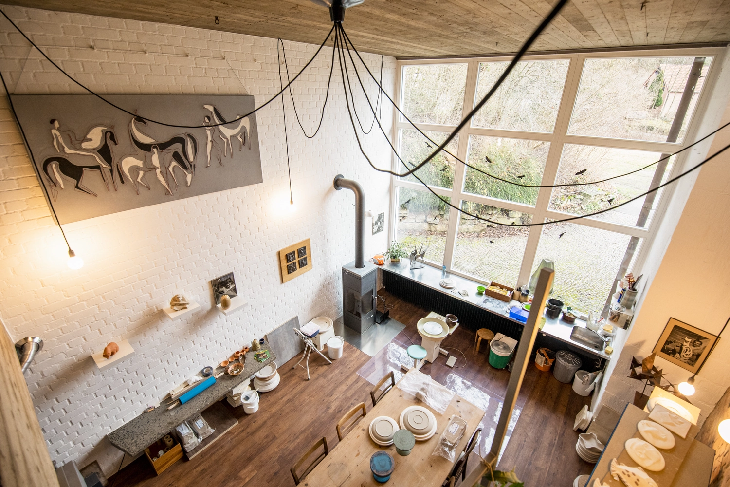 Keramik Atelier Melis - Töpferkurse - Ein erhöhter Blick auf einen sonnendurchfluteten, zweistöckigen offenen Wohnbereich mit Esstisch, Töpferwerkstatt, Holzofen und großen Fenstern.