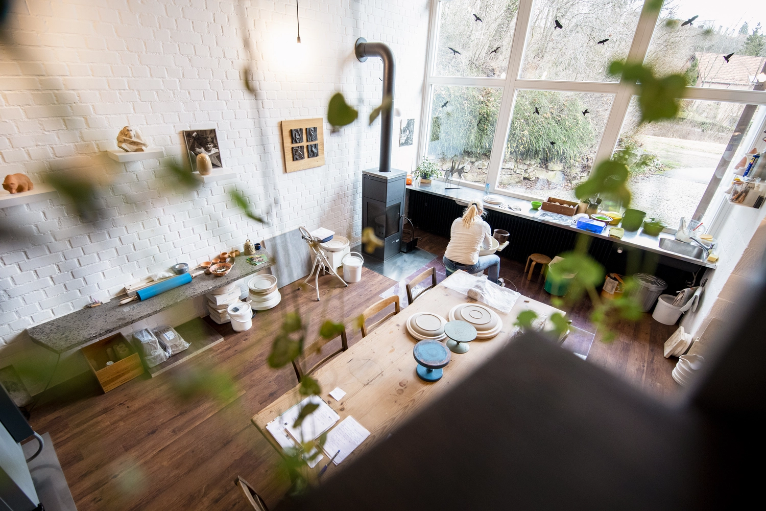 Keramik Atelier Melis - Töpferkurse - Draufsicht auf ein helles Töpferatelier mit einer Person, die an einem Tisch in der Nähe eines großen Fensters arbeitet, umgeben von Töpferwaren und Pflanzen.