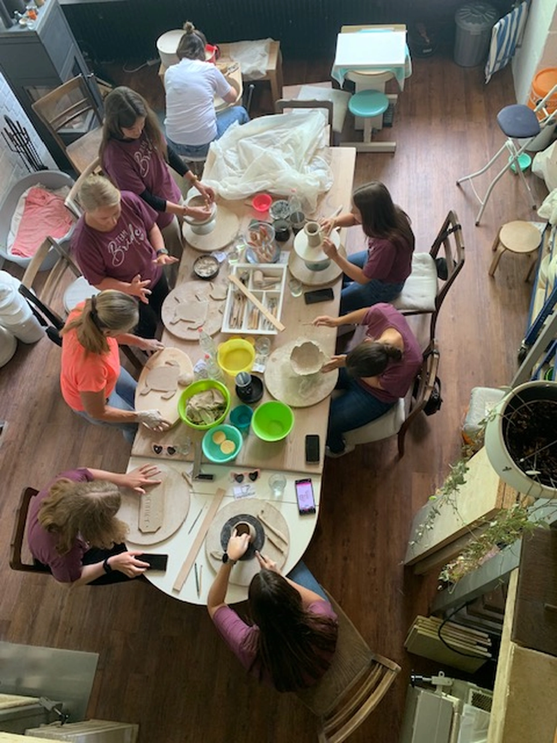 Keramik Atelier Melis - Töpferkurse - Eine Draufsicht auf eine Gruppe von Frauen, die in einem Töpferkurs in einem Atelier teilnehmen. Die Frauen sitzen um einen großen Holztisch und arbeiten an ihren Töpferscheiben oder formen Tonobjekte von Hand. Der Tisch ist mit verschiedenen Töpferwerkzeugen, Schalen und Tonresten bedeckt. Die Teilnehmerinnen tragen legere Kleidung, und der Atelierraum mit Holzboden und -möbeln strahlt eine warme, einladende Atmosphäre aus.